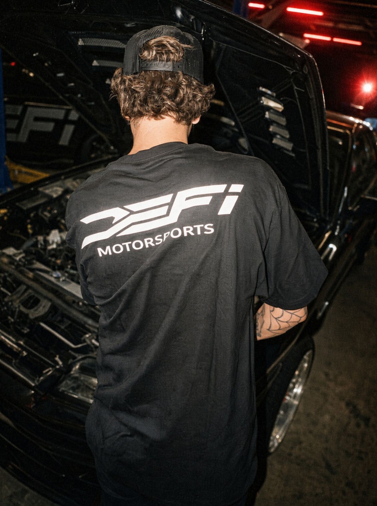 Wearing the black DEFI Motorsports t-shirt with the white back lockup, leaning into an engine bay